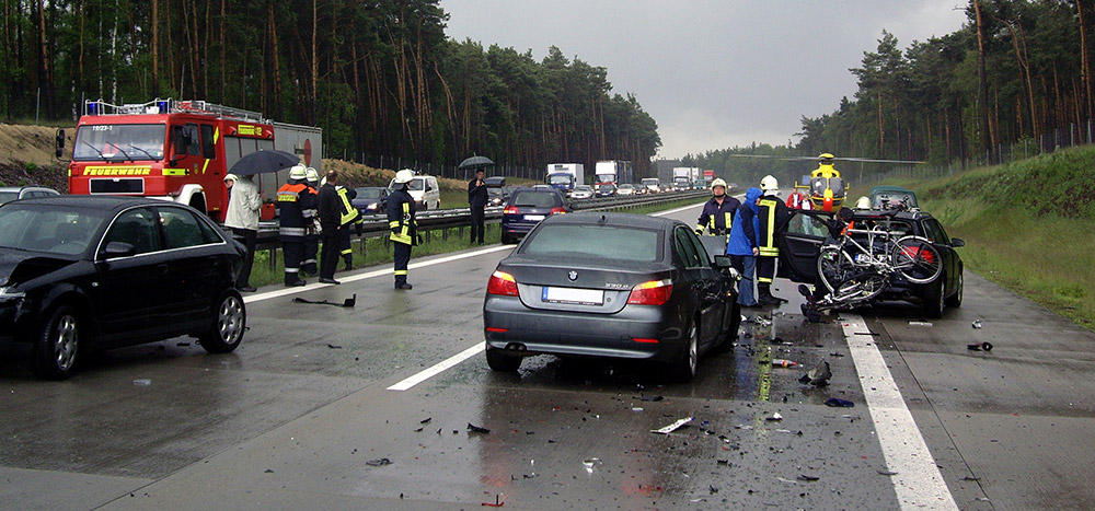 Der abgebildete Autobahnunfall mit Feuerwehreinsatz wird von Kfz Gutachtern begutachtet werden.  Der bereitstehende Rettungshubschrauber im Hintergrund weist auf einen Personenschaden hin.