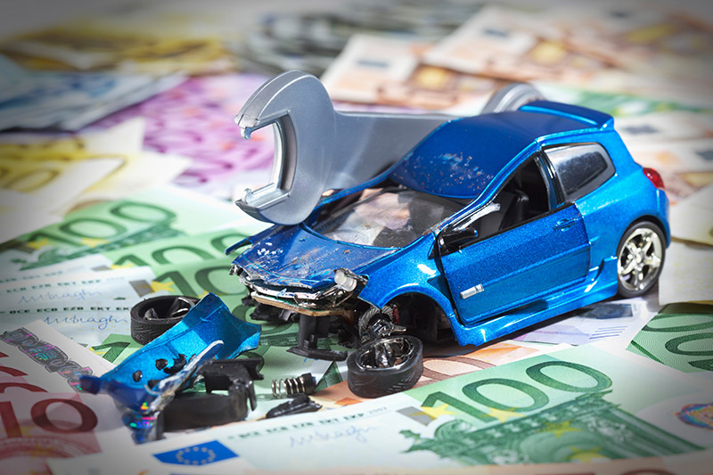 Ein Spielzeugauto mit zerstörter Front, auf der ein Schraubenschlüssel platziert ist, steht auf einem Teppich aus Geldscheinen.