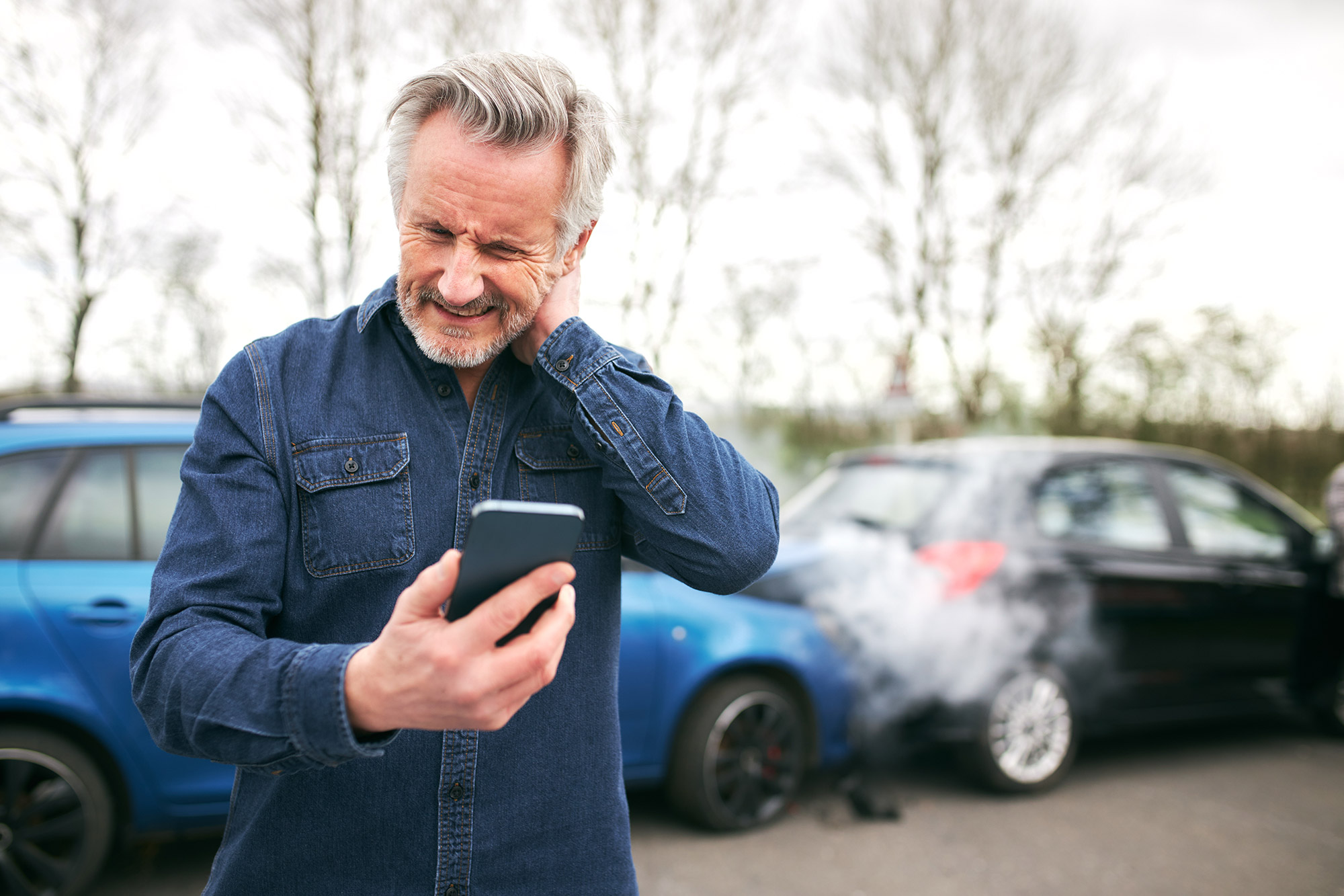 Diese Aufnahme zeigt einen Mann der, vor einem Unfall im Hintergrund, mit seinem Smartphone nach dem Hilfe Kontakt sucht.
